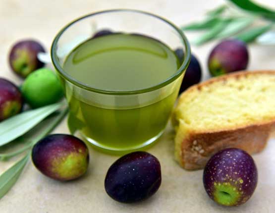 bicchiere-olio-evo bicchiere con olio di oliva e fetta di pane accanto, manciata di olive nere in primo piano e sullo sfondo